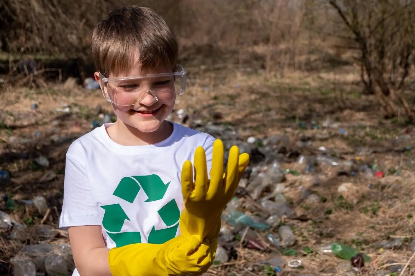 Junger weißer kaukasischer Junge mit einem Recycling-Symbol auf seinem T-Shirt und einer Brille, der gelbe Handschuhe anzieht.